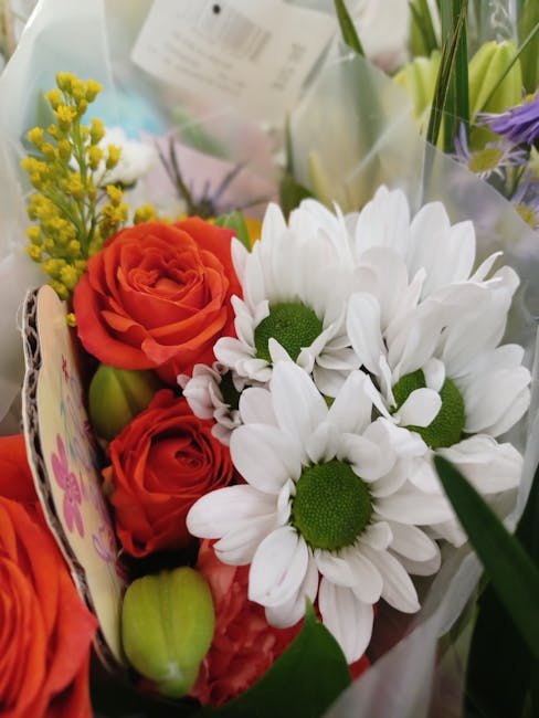 A close-up of a floral bouquet featuring two vibrant orange roses with tightly layered petals, surrounded by white daisies with large, rounded petals and bright green centers. The arrangement includes