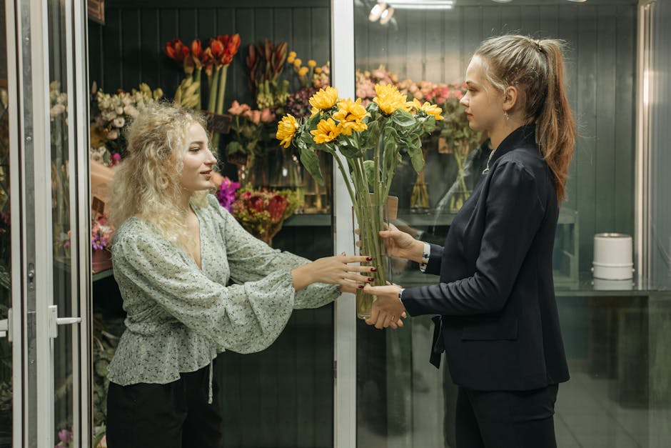 A florist with blonde curly hair, wearing a light-colored blouse with long sleeves, is receiving a bouquet of bright yellow sunflowers in a transparent glass vase from a customer with long brown hair 