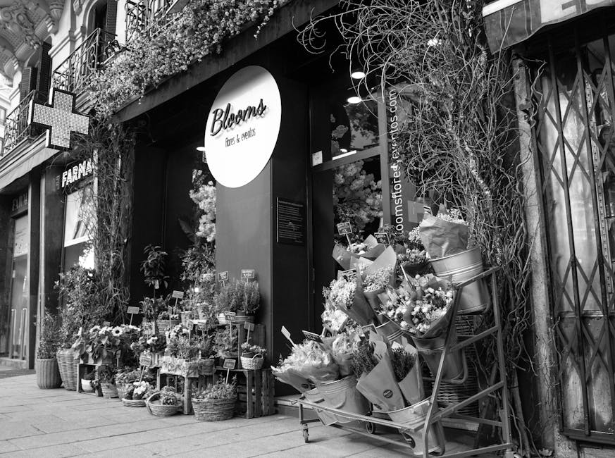 The exterior display of Bloom's flower shop, decorated with an array of vibrant fresh flowers including roses, lilies, daisies, and eucalyptus, arranged in baskets, bouquets, and vases placed on woode