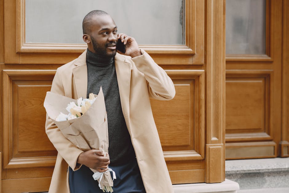 A man with dark skin, short hair, and a beard is standing outdoors in front of a wooden panel wall, holding a bouquet of fresh white roses wrapped in light brown paper. He is wearing a beige coat over