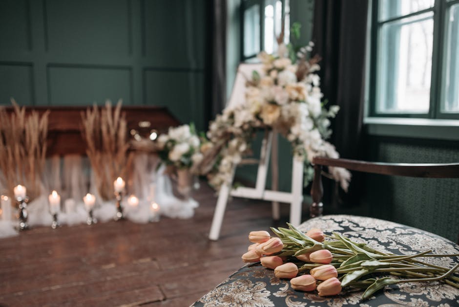 A floral arrangement set up in an indoor space with dark green paneled walls and wooden flooring. In the foreground, a bouquet of pale pink tulips with green stems rests on an ornate, upholstered cush