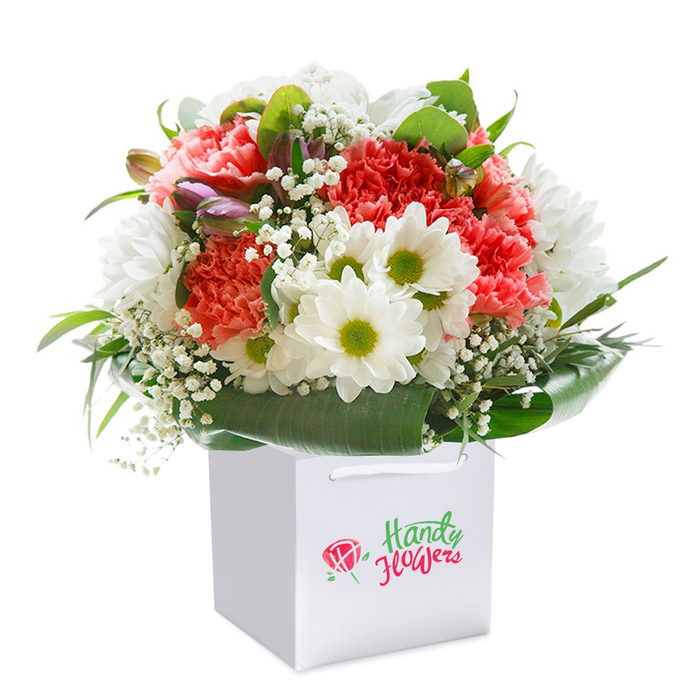 Standing tall within a modern white gift bag, this meticulously arranged bouquet sings with colour and texture-an embodiment of Bracknell Forest's floral artistry. The focal point is an inviting mix of coral, peach, and radiant red carnations, their frilled, abundant petals layered in plush clouds. White daisy-like chrysanthemums, each with a bright chartreuse centre, dot the ensemble, imparting cheer and clarity reminiscent of the wild blooms at Englemere Pond or the family-friendly gardens in Owlsmoor. Delicate clouds of gypsophila interlace among the larger blossoms, lending an airy, cloud-soft finish, while discreet purple-tinted buds, most likely alstroemeria, evoke anticipation and depth in the left pocket of the bouquet. The whole arrangement is ringed by a lush coronet of deep green, glossy leaves, mirroring the tranquil canopies found in Bracknell Forest's woodlands. The stylish gift bag, with sturdy white ribbon handles, delivers practical elegance for occasions ranging from local anniversaries to spontaneous doorstep surprises in Blackwater or Bagshot. Each element is lit suavely against a crisp white background, amplifying the freshness and high standard expected from Bracknell Forest florists. The result is a balanced, rounded bouquet that radiates warmth, refinement, and heartfelt intent-ideal for birthdays, important events, or simply sharing a slice of the area's natural beauty.